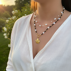 Person wearing a pearl and black beaded necklace with a gold pendant in a natural setting.